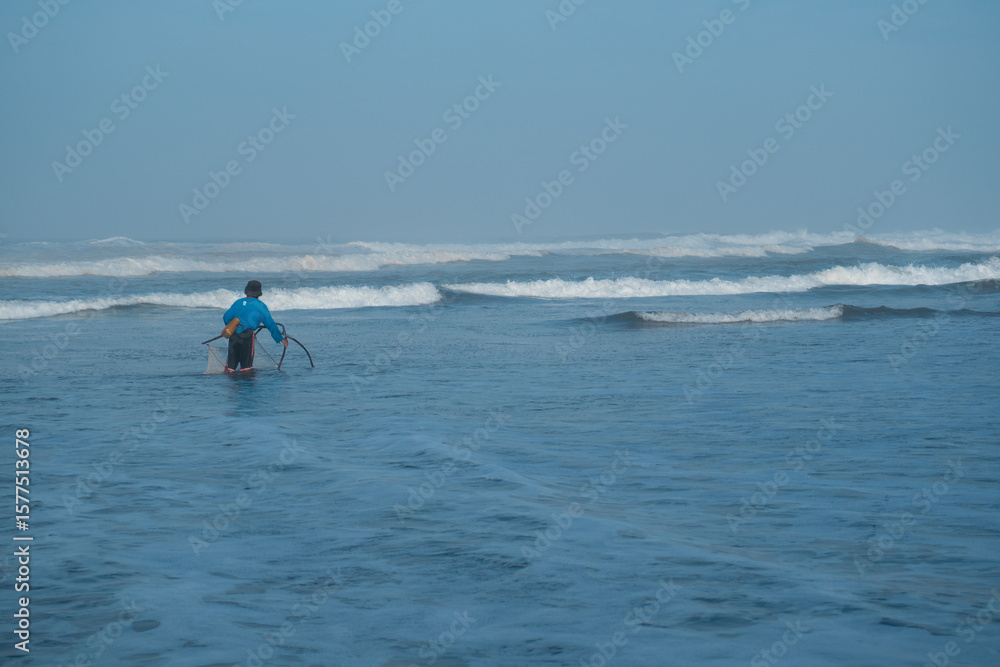 Naklejka premium Yogyakarta, Indonesia-July 3, 2022: A lone fisherman pulling a net in the shallow ocean waters at the beach, under a vast sky, depicting traditional livelihood.