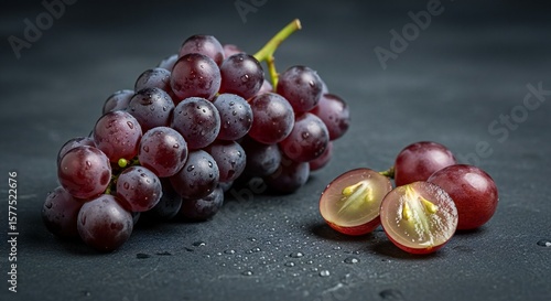A fresh bunch of crimson grapes with water droplets and sliced fruit on a dark background.