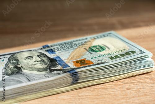 Stack of one hundred dollar banknotes on wooden desk in warm sunlight with selective focus, concept of cash payment, wealth, savings, investment, financial success and money background.