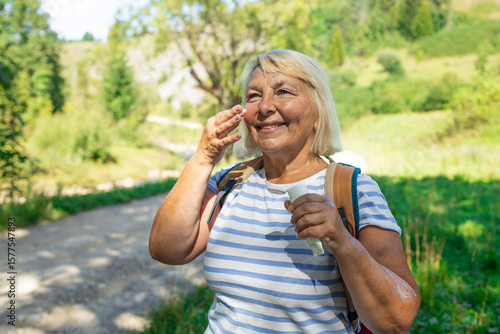 A cheerful senior woman standing on a scenic forest trail applies face cream or sunscreen while enjoying the outdoors. Wearing a striped shirt and backpack, she symbolizes active aging