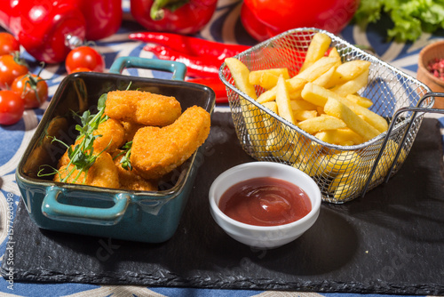 The photo shows a black slate serving board with a teal dish containing breaded chicken nuggets, garnished with microgreens. Next to it is a metal basket filled with golden French fries. 
