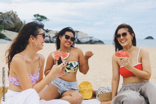 Three happy young Asian women enjoying pizza and drinks during a beach picnic on a sunny summer day. Group of friends relaxing on vacation, laughing and having fun outdoors near the sea.