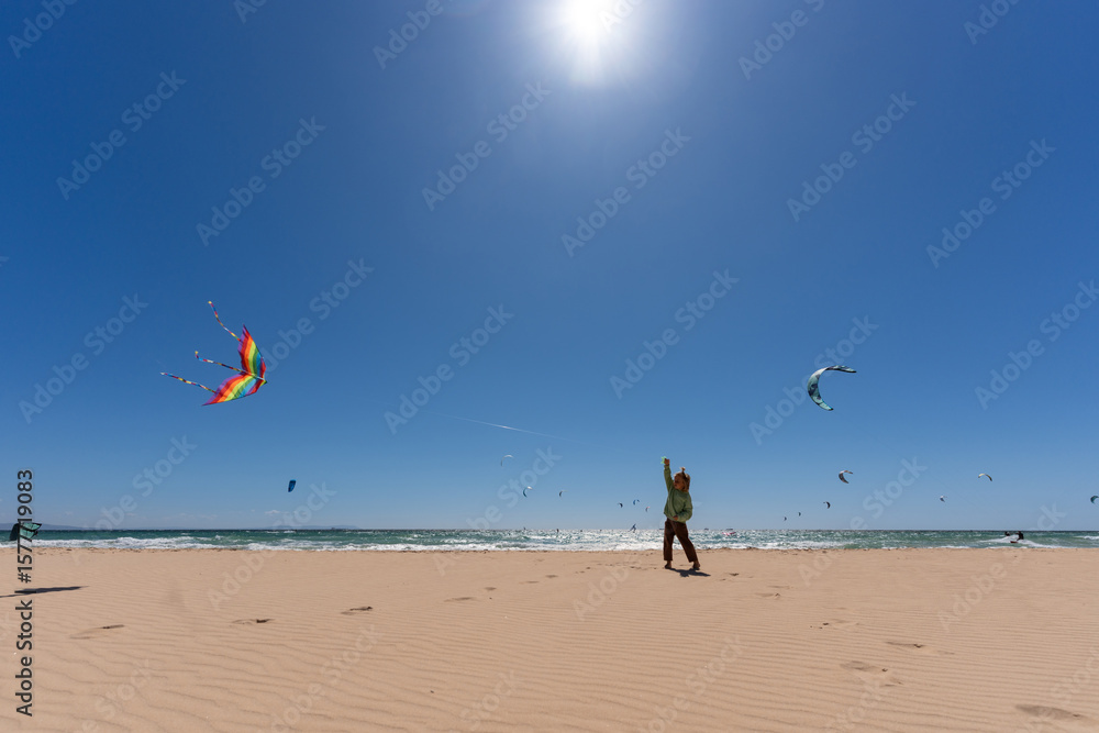 Obraz premium Young child flying a colorful rainbow kite on a sunny beach with a clear blue sky and kitesurfers in the background. Concept of summer fun, childhood joy, and outdoor freedom
