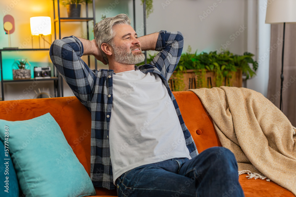 Fototapeta premium Tired senior man with gray hair in casual clothes stretching arms and resting on sofa in living room. Thoughtful Caucasian middle-aged guy relaxing on couch with hands behind head looking away at home