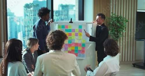 Diverse group of professionals discussing ideas with sticky notes on a whiteboard in a bright, modern office meeting room.