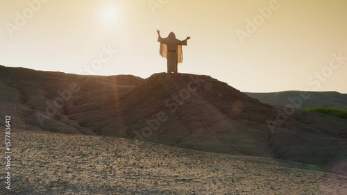 Jesus standing in the wilderness at sunset, raising His hands in prayer under�the�open�sky. Extreme Long Shot