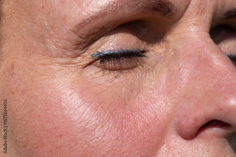 Obraz premium Macro photo of a closed eye showing realistic wrinkles, natural pores, and mature skin tone in sunlight. Useful for skincare, dermatology, or aging-focused creative projects.