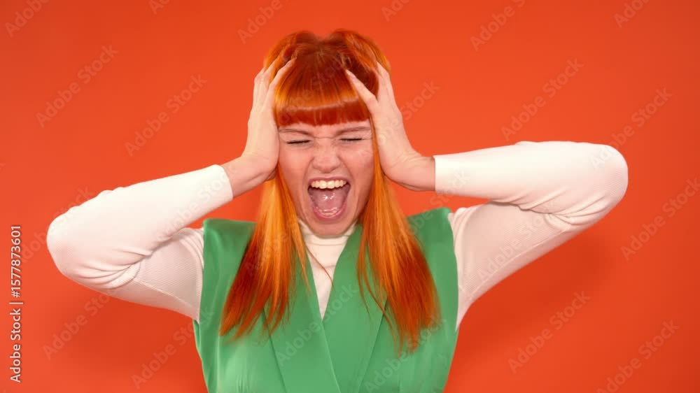 Young woman with orange and red hair expressing frustration or anger by holding her head with both hands She wearing green vest over white long-sleeve shirt, set against orange background Young woman with orange and red hair expressing frustration or anger by holding her head with both hands She wearing green vest over white long-sleeve shirt, set against orange background