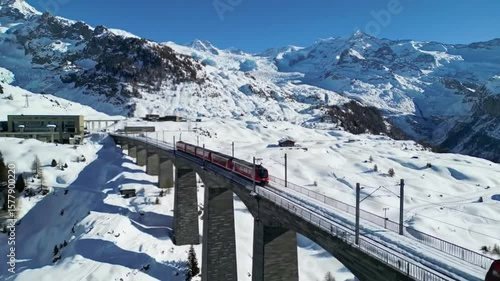Red train crossing a viaduct in a snowy mountain landscape on a clear and sunny winter day