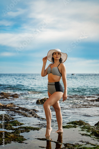 Elegant Woman in Retro Swimsuit on Rocky Coastline
A woman in a polka-dot bikini and white sun hat stands gracefully on a rugged, wet rocky beach, with the ocean and cliffs in the background