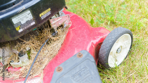 A detailed close-up of a red lawn mower set against a grassy backdrop, focusing on the wheel and undercarriage showing accumulated grass. The image captures the practical aspect of garden care equipme
