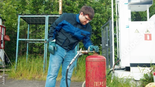 A man in a blue uniform tightens the valve of a red propane tank and disconnects the nozzle. Refueling with liquefied gas at a station on a summer day. Transportation of natural gas for domestic use