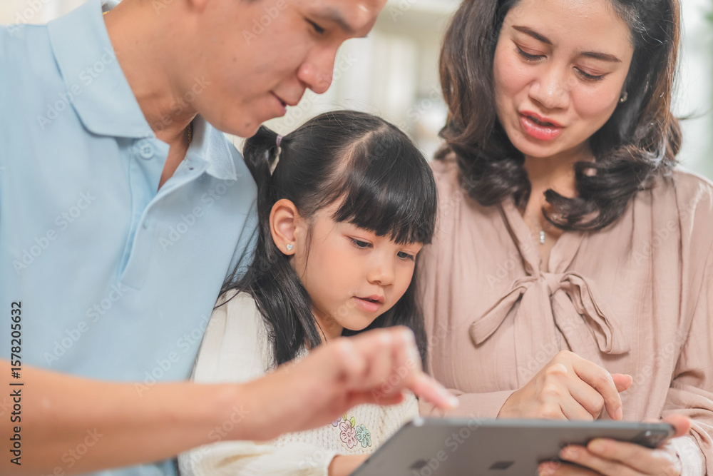 Fototapeta premium Happy Asian family with father mother and daughter using digital tablet laptop for home-based learning education while watching movies cartoons sitting together on living room sofa creating joyful