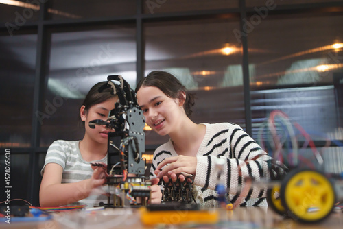 Two girl students, Asian and Caucasian, working together on building a robotic arm for a STEM project in class, focusing and smiling while connecting parts.