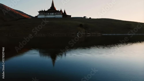 a tranquil and scenic view of a traditional Asian-style temple situated on a grassy hilltop, reflected beautifully in the still waters of a lake below.