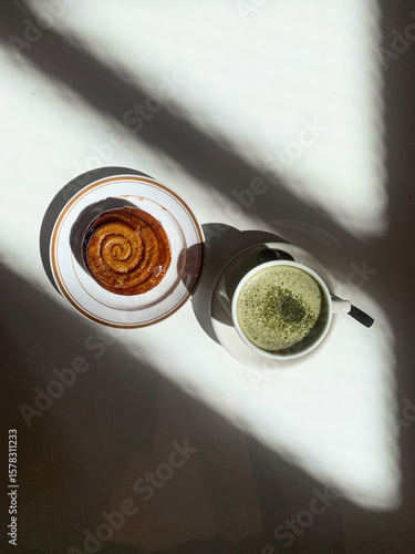 Golden cinnamon roll with glossy glaze served on a plate beside a creamy matcha latte. Bright sunlight casts artistic shadows on the white table.