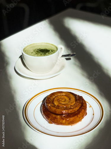 Golden cinnamon roll with glossy glaze served on a plate beside a creamy matcha latte. Bright sunlight casts artistic shadows on the white table.