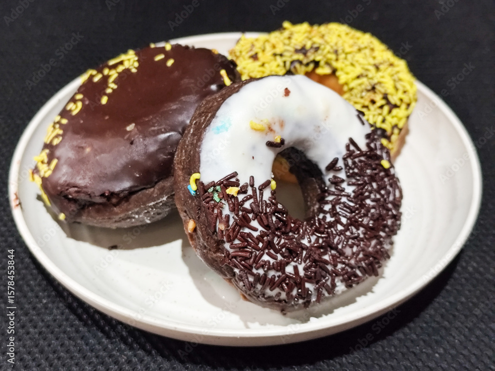 Fototapeta premium Three delicious donuts topped with sprinkles and various colored glazes, displaying a tempting texture, served on a white plate.