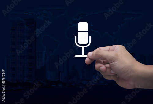 Fototapeta Naklejka Na Ścianę i Meble -  Hand holding microphone flat icon in fingers over world map, modern city tower and skyscraper, Technology communication concept, Elements of this image furnished by NASA