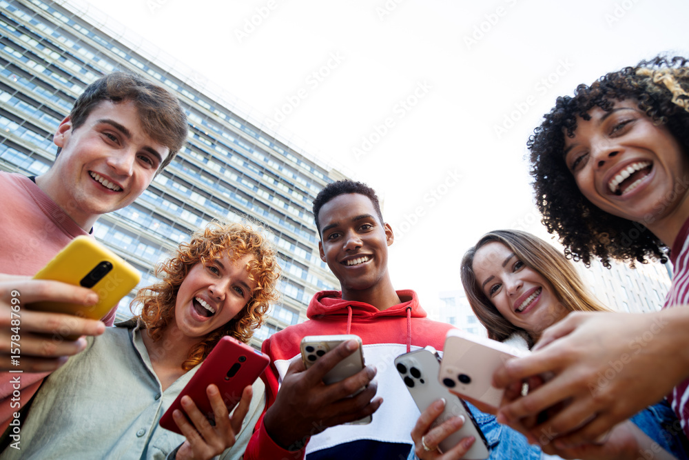 Obraz premium Happy students using smartphones in university campus: low angle view of cheerful diverse friends enjoying mobile technology