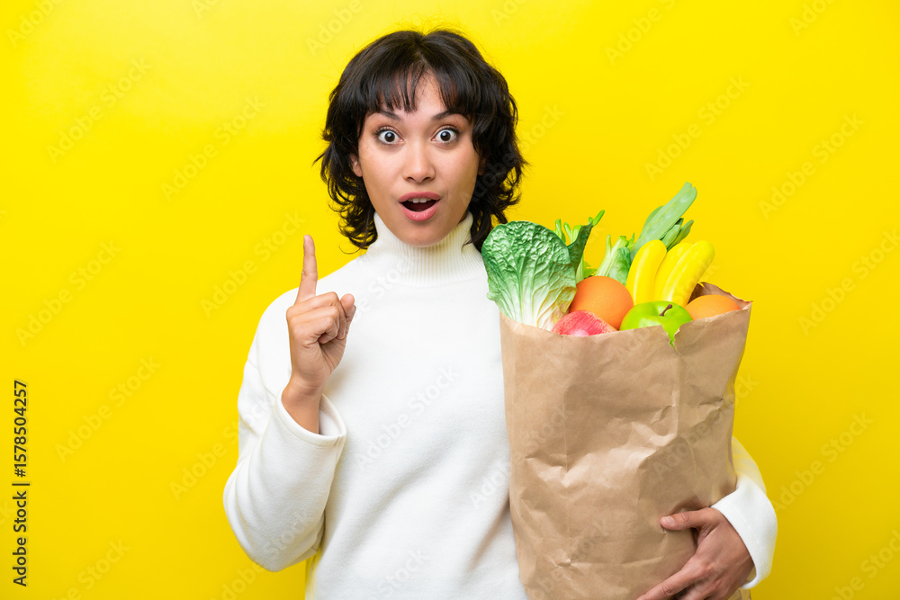Young Argentinian woman holding a grocery shopping bag isolated on yellow background intending to realizes the solution while lifting a finger up Young Argentinian woman holding a grocery shopping bag isolated on yellow background intending to realizes the solution while lifting a finger up