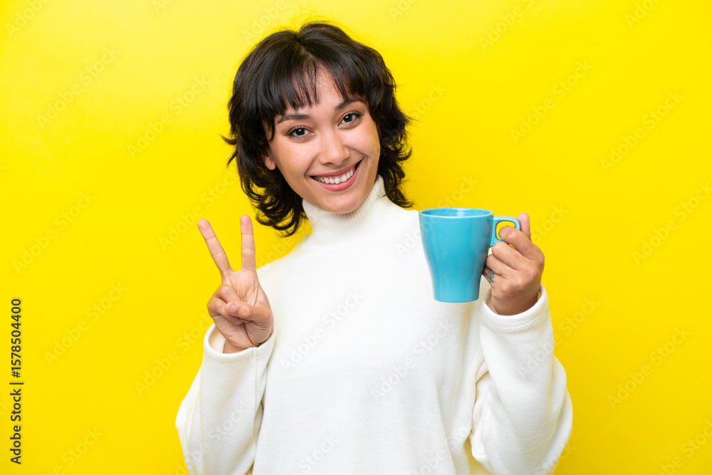 Young Argentinian woman holding cup of coffee isolated on yellow background smiling and showing victory sign Young Argentinian woman holding cup of coffee isolated on yellow background smiling and showing victory sign