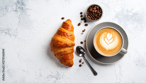 Minimalist flat lay of a fresh croissant and a latte with beautiful foam art on a light-textured stone background.