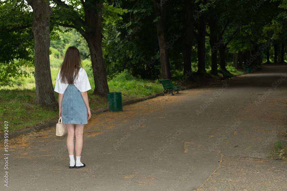 A young woman dressed in a casual outfit pauses on a quiet park path. Sunlight filters through the trees, creating a serene atmosphere. She seems contemplative, enjoying the solitude A young woman dressed in a casual outfit pauses on a quiet park path. Sunlight filters through the trees, creating a serene atmosphere. She seems contemplative, enjoying the solitude