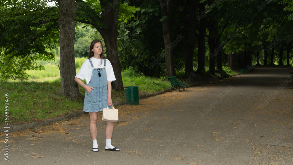 Fototapeta premium A young woman stands confidently in denim overalls, holding a small bag, surrounded by lush greenery in a peaceful park during the day. She enjoys the warm sun and fresh air