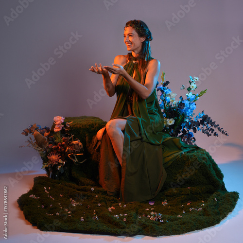 fantasy portrait beautiful female model wearing green fairy fantasy gown costume. Graceful sitting pose, moss chair with floral nature decor. Silhouetted figure of person, colourful studio background.