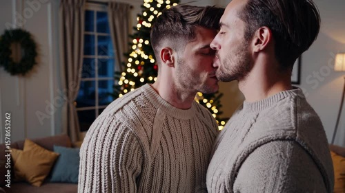 Romantic gay couple kissing in cozy home by Christmas tree wearing knit sweaters