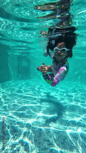 A young girl stands still in a swimming pool, looking directly at the camera with a serene expression.