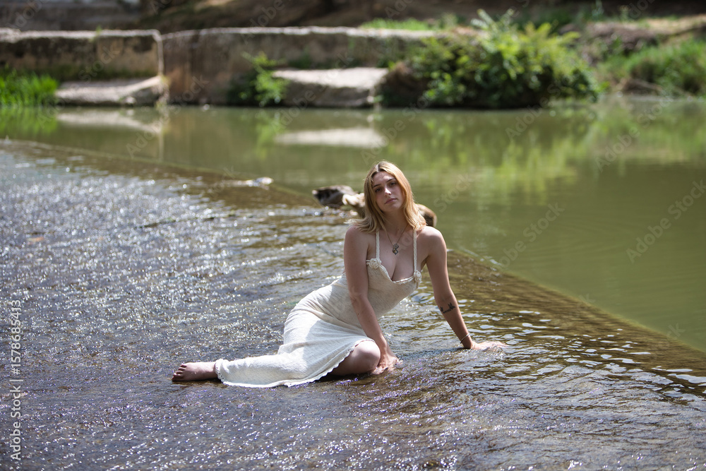 Fototapeta premium A young, beautiful blonde woman wearing a beige knitted dress sits by a small waterfall on a hot summer's day in Spain. There are ducks and vegetation in this unrivalled natural setting.