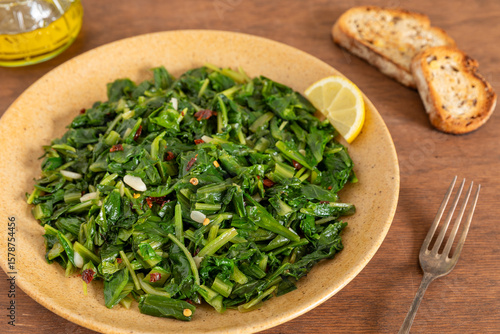 Roman-style chicory greens sautéed in olive oil (Cicoria Ripassata alla Romana) - a traditional Italian side dish served with toasted bread and a slice of lemon. Brown wooden table.