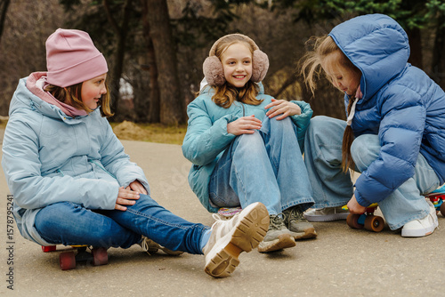 Teen girls in jackets, jeans sit on skateboards in city park. Winter street sport creates fun, friendship, confidence, freedom. Outdoor activity builds childhood energy, motion, lifestyle, expression