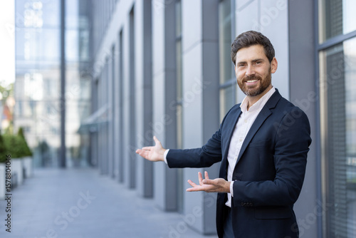 Принти на полотні A businessman in a suit gestures with both hands, welcoming or presenting something in front of a modern building