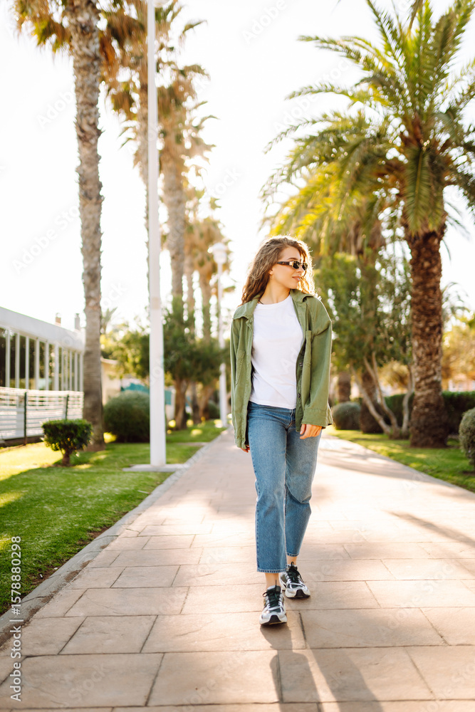 Cheerful woman with curly hair and sunglasses enjoys a sunny day on an alley with palm trees. A beautiful tourist with a backpack is walking outdoors. Leisure, weekend concept. Cheerful woman with curly hair and sunglasses enjoys a sunny day on an alley with palm trees. A beautiful tourist with a backpack is walking outdoors. Leisure, weekend concept.