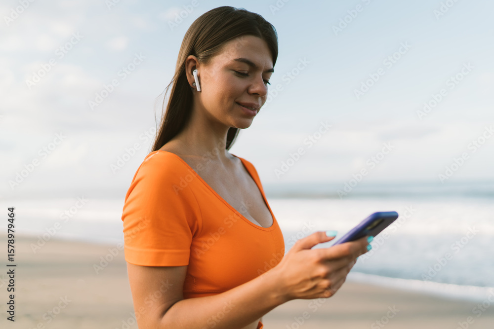 Naklejka premium Young woman in bright outfit focused on smartphone screen while wearing wireless earbuds, symbolizing digital lifestyle, tech immersion and mobile connectivity in outdoor freedom.