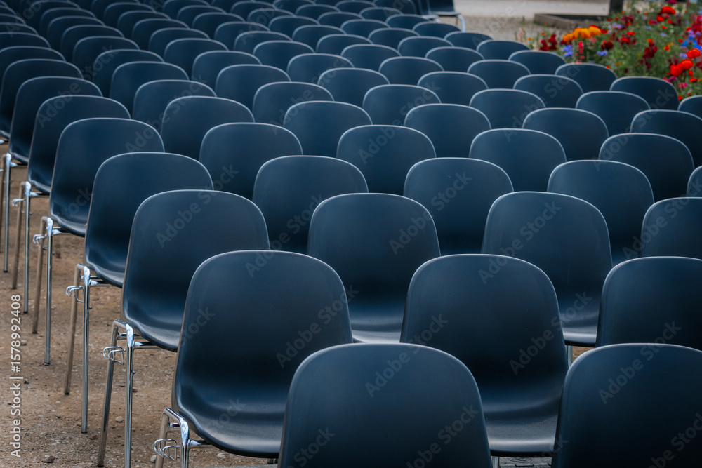Fototapeta premium Multiple rows of identical black chairs are neatly arranged in an outdoor setting on a dirt surface, creating a uniform and symmetrical pattern.