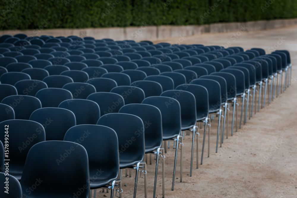 Fototapeta premium Multiple rows of identical black chairs are neatly arranged in an outdoor setting on a dirt surface, creating a uniform and symmetrical pattern.