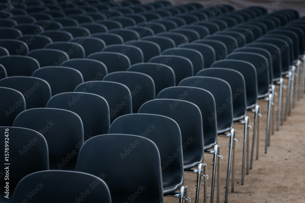 Fototapeta premium Multiple rows of identical black chairs are neatly arranged in an outdoor setting on a dirt surface, creating a uniform and symmetrical pattern.