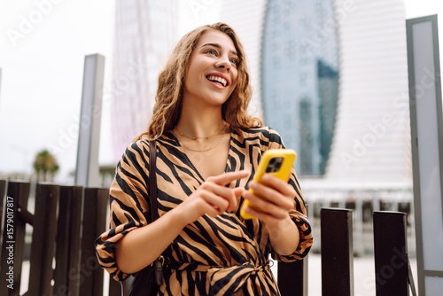 Fotografie Smiling woman with a phone in her hands against the backdrop of the bustle of the city