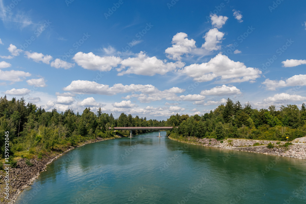 Fototapeta premium Turquoise river flowing under a bridge, surrounded by lush green forest and blue sky with clouds. Scenic nature landscape perfect for travel, outdoor, and environmental themes