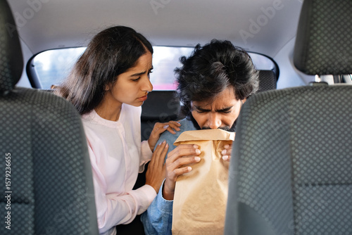 Fotografi Worried Indian woman comforting man vomiting into paper bag in backseat