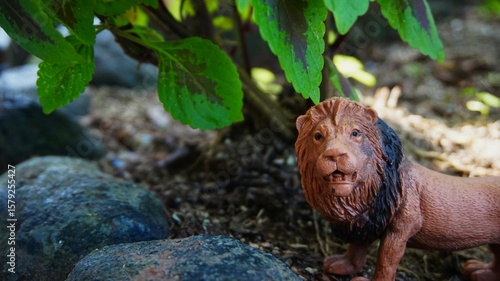 Lion toy, close up of a brown rubber lion toy with a background of plants, soil and rocks
