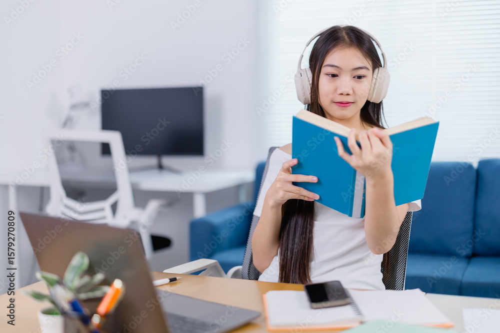 Concentrated young asian female student reading a book and listening to music with headphones while sitting at desk and studying online from home, elearning concept Concentrated young asian female student reading a book and listening to music with headphones while sitting at desk and studying online from home, elearning concept