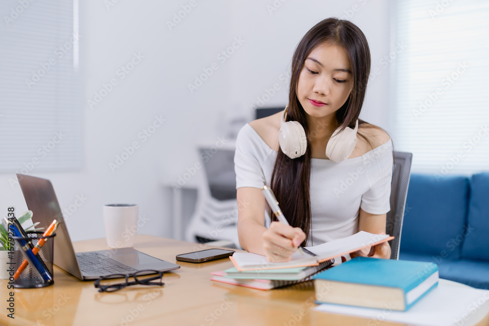 Focused young Asian woman wearing headphones around her neck is taking notes and studying with laptop, books, and smartphone on a desk at home Focused young Asian woman wearing headphones around her neck is taking notes and studying with laptop, books, and smartphone on a desk at home