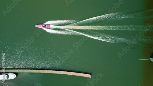 Top-down aerial shot of a pink speedboat leaving a V-shaped wake on green water. Dynamic summer scene captured by drone.