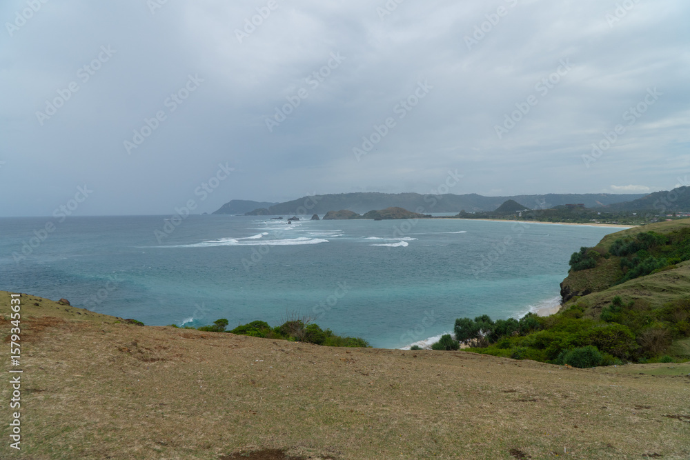 Obraz premium Scenic ocean view in Lombok, Indonesia. Waves crash on rocky islets with turquoise water and lush coastline under a cloudy sky