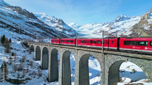 Red train crossing a stone bridge in a snowy mountain landscape on a clear and sunny winter day